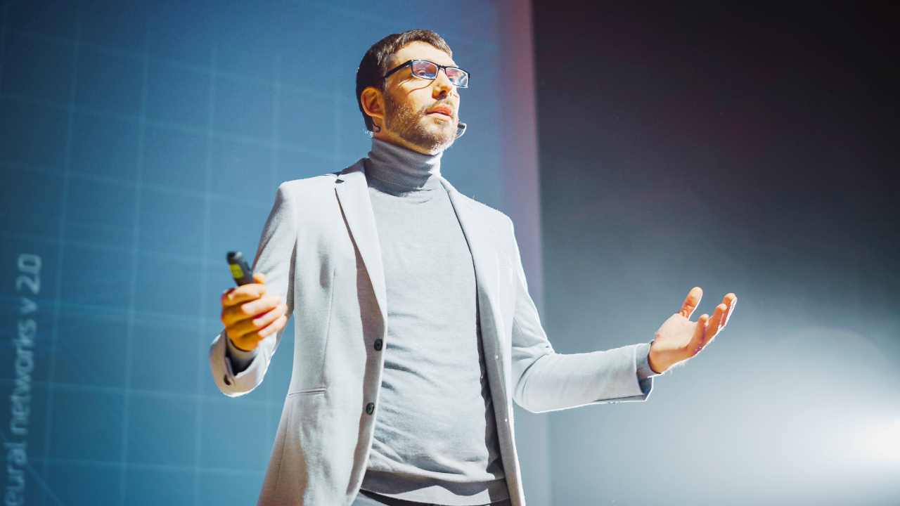 Here is an alt text description for the image: "A professional speaker wearing a light-colored blazer and turtleneck is presenting on stage. He is holding a presentation remote in one hand and gesturing with the other. He has a beard, glasses, and a confident expression. The stage is illuminated with bright lighting, and a large screen in the background displays faint text and graphics related to technology or neural networks."