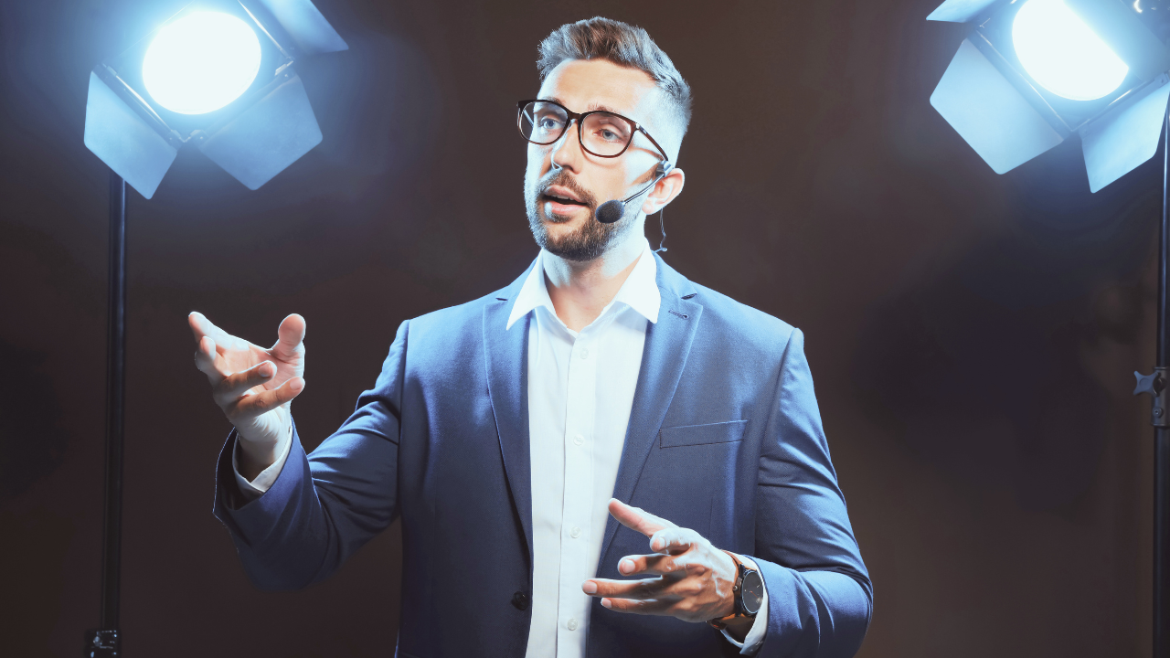 A professional speaker wearing a navy blue suit, white dress shirt, and glasses, delivering a presentation on stage. He is using a headset microphone and gesturing with his hands while speaking. Bright stage lights shine behind him, illuminating the scene.