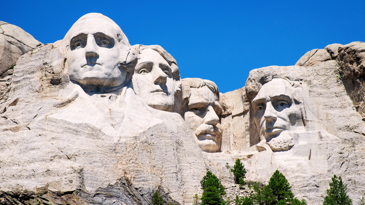 Mount Rushmore National Memorial in South Dakota on a clear day, featuring the carved faces of George Washington, Thomas Jefferson, Theodore Roosevelt, and Abraham Lincoln against a bright blue sky, surrounded by rocky terrain and green trees.