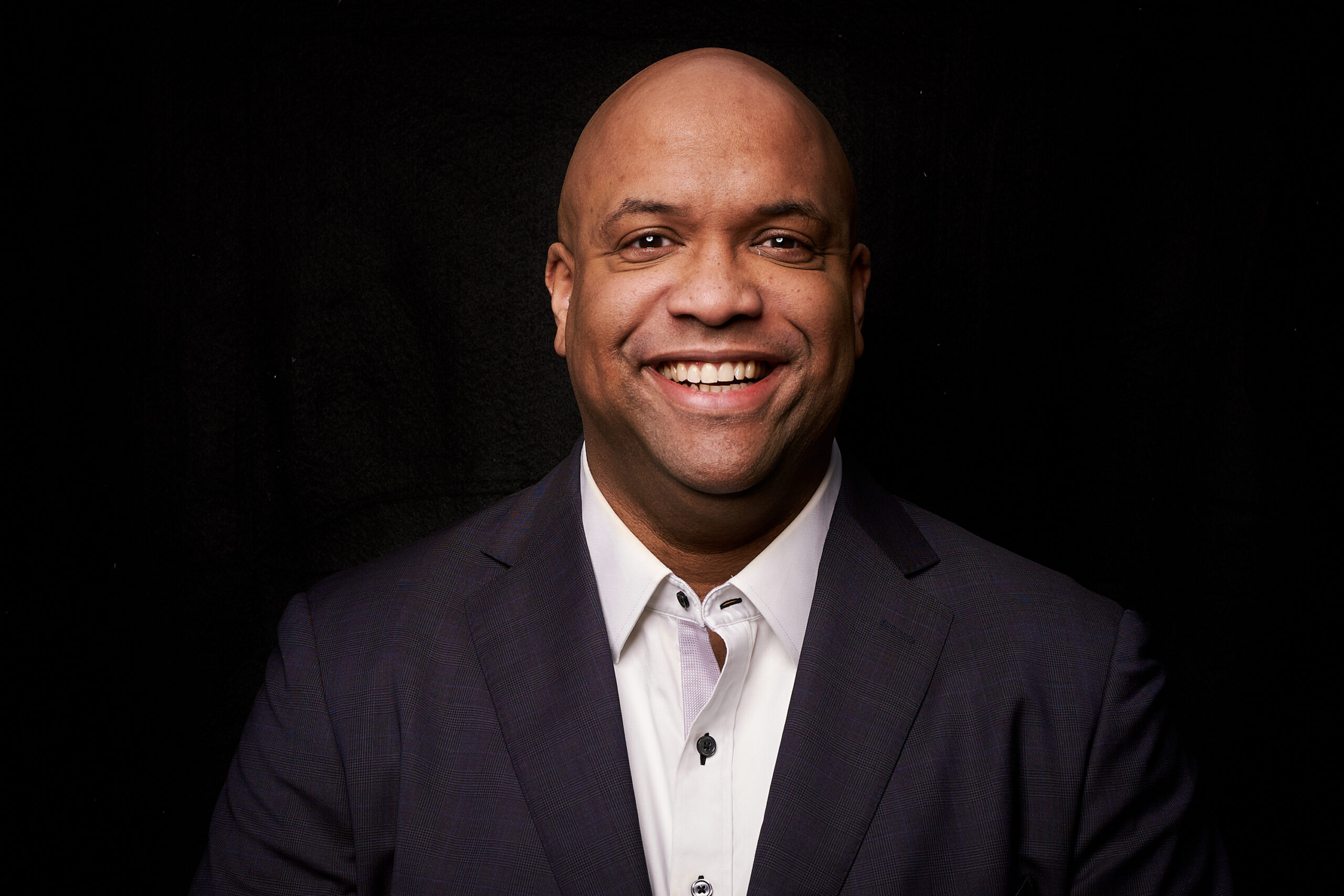 Mike Veny, a bald black man, is wearing a dark suit and a white dress shirt. He stands against a black background, smiling warmly and confidently at the camera.