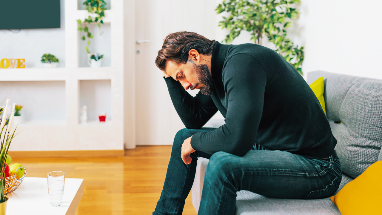 Alt text: A man with dark hair, a beard, and a black long-sleeve shirt sits on a gray couch in a modern living room. He rests his elbow on his knee and holds his head in his hand, appearing distressed or deep in thought. The room has white walls, built-in shelves with plants and decorative items, and yellow accent pillows on the couch. A small table in the foreground holds a glass of water and a fruit basket.