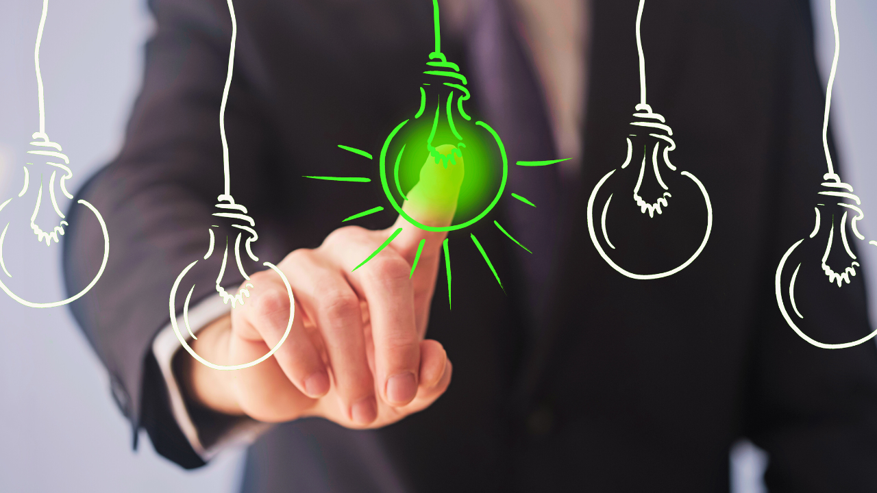 A business professional in a suit is pointing at a glowing green light bulb, surrounded by white hand-drawn light bulbs. The image symbolizes innovation, creativity, and new ideas.