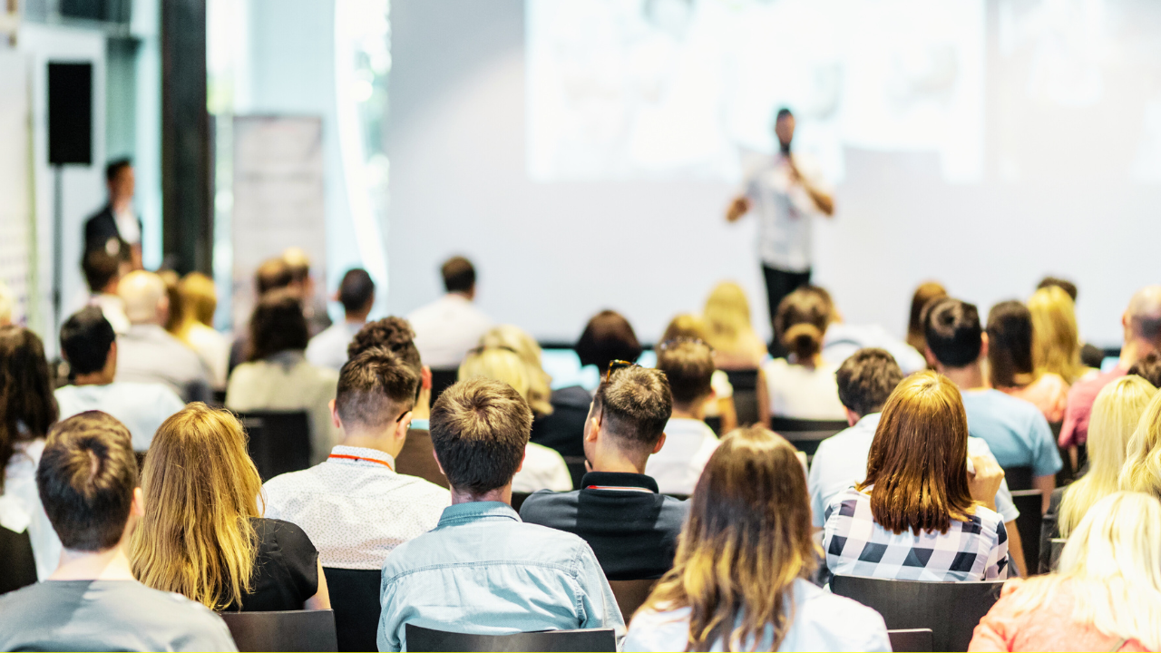 A group of people seated and facing a speaker on stage during a professional conference or seminar. The speaker is standing in front of a large screen, presenting to the audience in a well-lit, modern conference room.