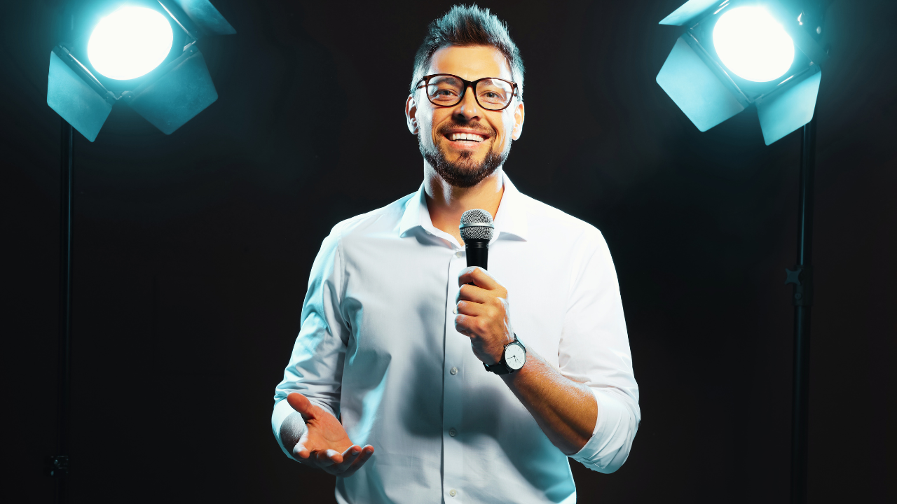 A smiling man wearing glasses and a white shirt holds a microphone while speaking on stage, standing under bright studio lights — representing engaging mental health speakers who inspire audiences.