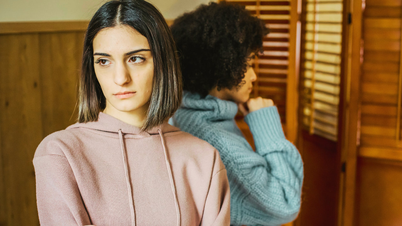 Two women stand back-to-back in a tense, silent moment after an argument. The woman in front, wearing a pink hoodie, looks away with a mix of sadness and frustration, while the woman behind her in a blue sweater stares down, appearing deep in thought. The warm, wooden background suggests an intimate setting where emotions still linger. This image visually represents the struggle of forgiveness, resentment, and emotional distance—capturing the theme of how to forgive when you’re still angry.