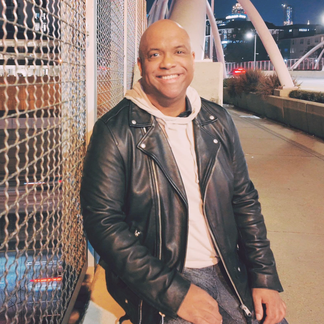 Mike Veny smiling confidently on a city bridge at night, dressed in a black leather jacket and light hoodie, with glowing streetlights, traffic below, and modern architectural arches framing the urban skyline.