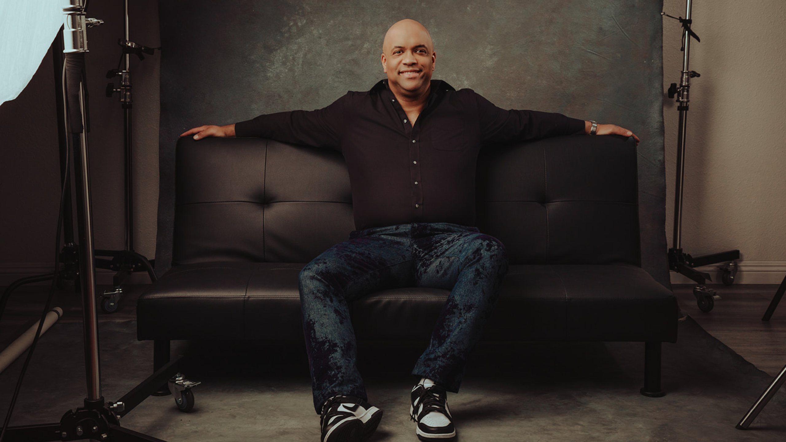 Mike Veny, a bald, smiling male mental health keynote speaker, sits confidently on a black leather couch in a professional studio setting, arms stretched across the backrest. He wears a fitted black button-down shirt, dark patterned jeans, and black-and-white sneakers. The background features a textured gray backdrop with visible studio lighting equipment on both sides, creating a polished, high-contrast portrait that conveys confidence, approachability, and professionalism.