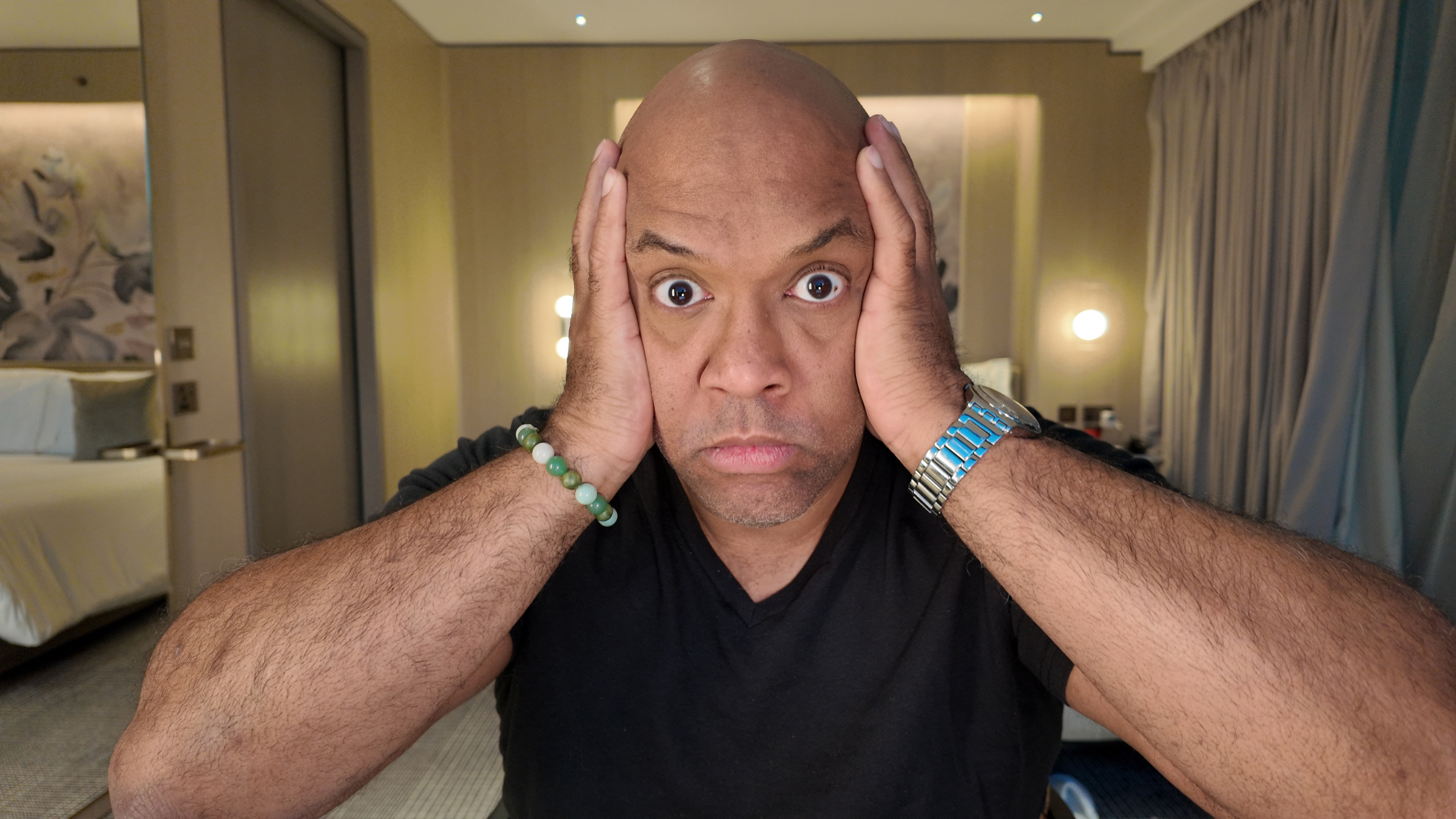 Mike Veny, a workplace mental health speaker, looks directly at the camera with wide eyes and hands pressed to his face, expressing stress and overwhelm in a modern hotel room setting, illustrating the emotional challenges professionals face in today’s workplace.