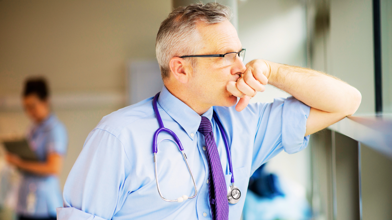 Middle-aged male healthcare professional wearing glasses, a light blue dress shirt, and a purple tie, with a stethoscope around his neck, stands by a hospital window looking outside with his hand resting against his mouth in a thoughtful, concerned expression; a blurred nurse using a tablet appears in the background, suggesting a busy clinical environment and themes of healthcare stress, decision-making, and leadership in medical settings.