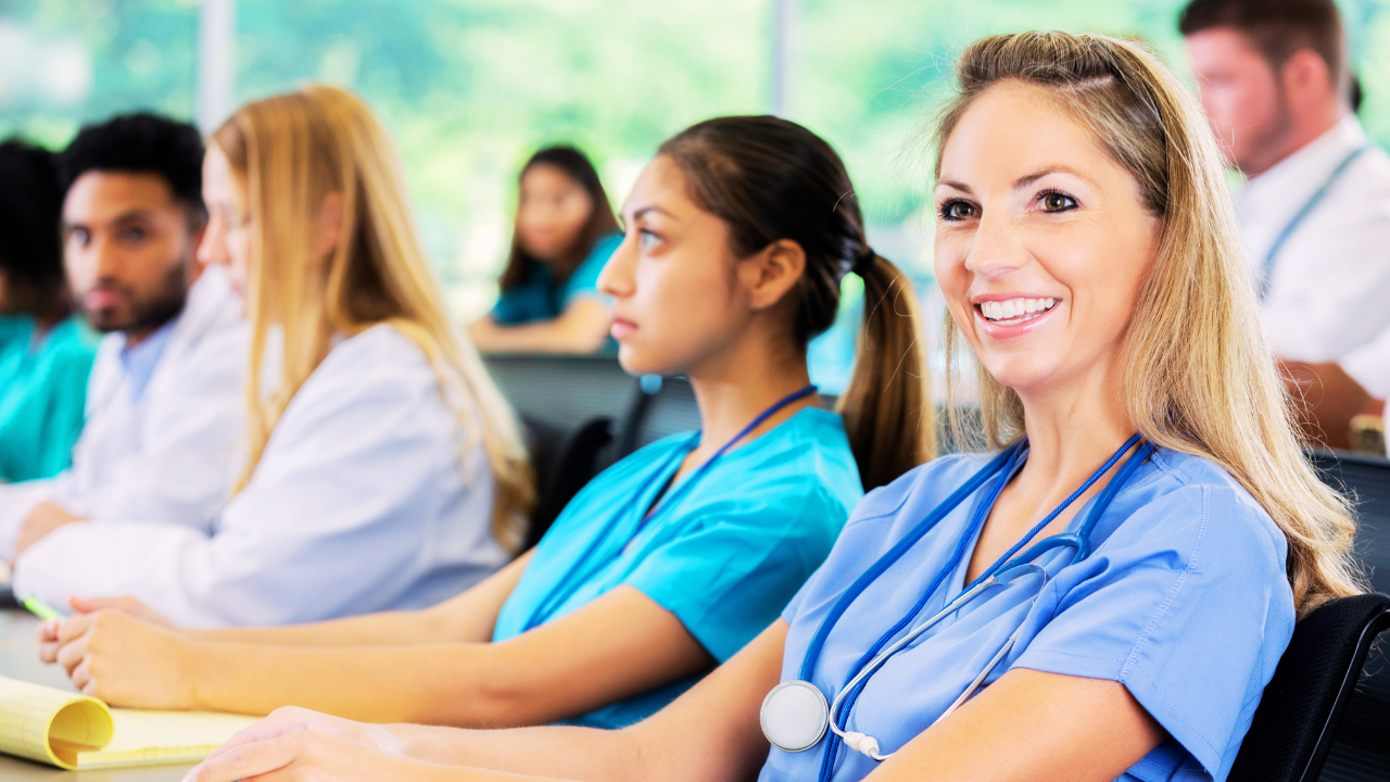 A diverse group of healthcare professionals in scrubs attend a professional training session or conference workshop, listening attentively in a classroom-style setting. A smiling nurse in the foreground wears a stethoscope and looks engaged, while colleagues take notes beside her. The scene represents continuing education, CEU opportunities, and professional development at a live event or keynote presentation.