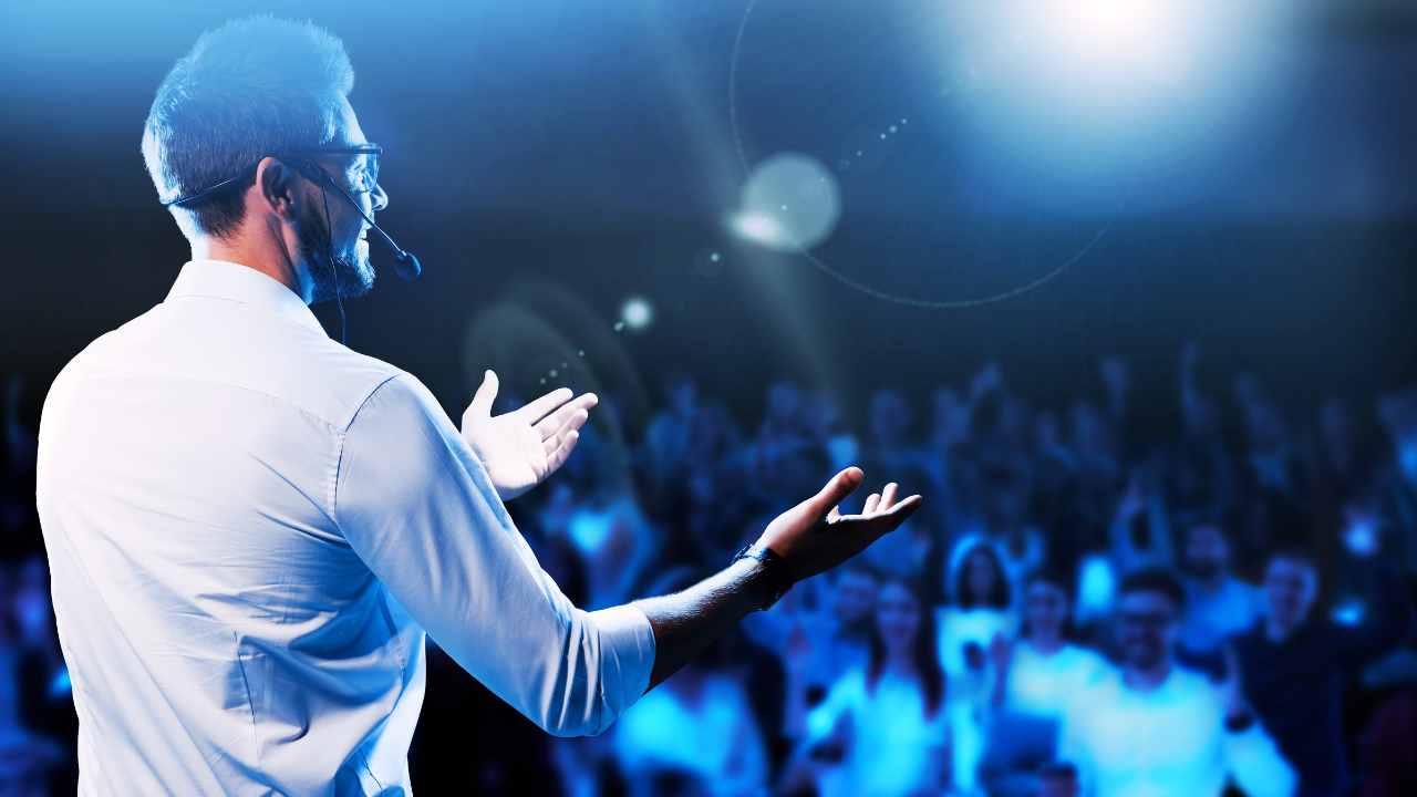 Mental health keynote speaker delivering an engaging presentation on stage, viewed from behind and slightly to the side. The speaker, a man wearing a fitted white shirt and a headset microphone, gestures with open hands toward a large audience. Bright stage lights create a blue-toned atmosphere with lens flares, highlighting the energy of the room. The audience appears attentive and inspired, slightly blurred to emphasize the speaker’s presence and authority. The scene conveys connection, confidence, and impact, illustrating a dynamic mental health keynote speaker leading a powerful, interactive talk on emotional well-being and resilience.
