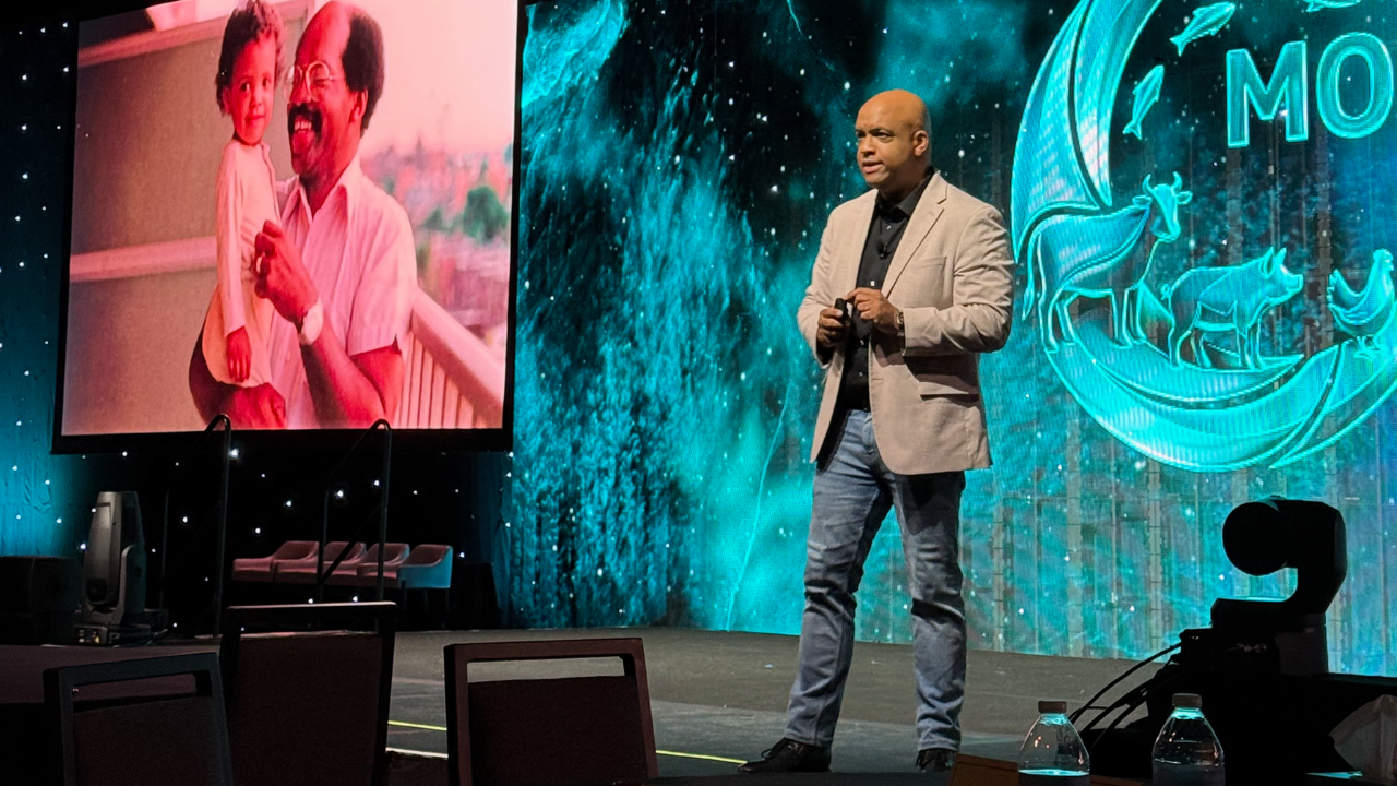 Global Meetings Industry Day – GMID Mike Veny motivational speaker delivering a keynote at a corporate meeting for Global Meetings Industry Day (GMID), standing on stage with teal lighting, large presentation screen, and professional conference environment.