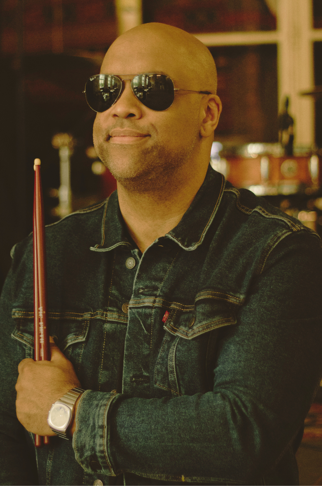 Mike Veny drummer and mental health speaker wearing sunglasses and a denim jacket, holding drumsticks in a music studio setting, representing resilience, creativity, and emotional expression.