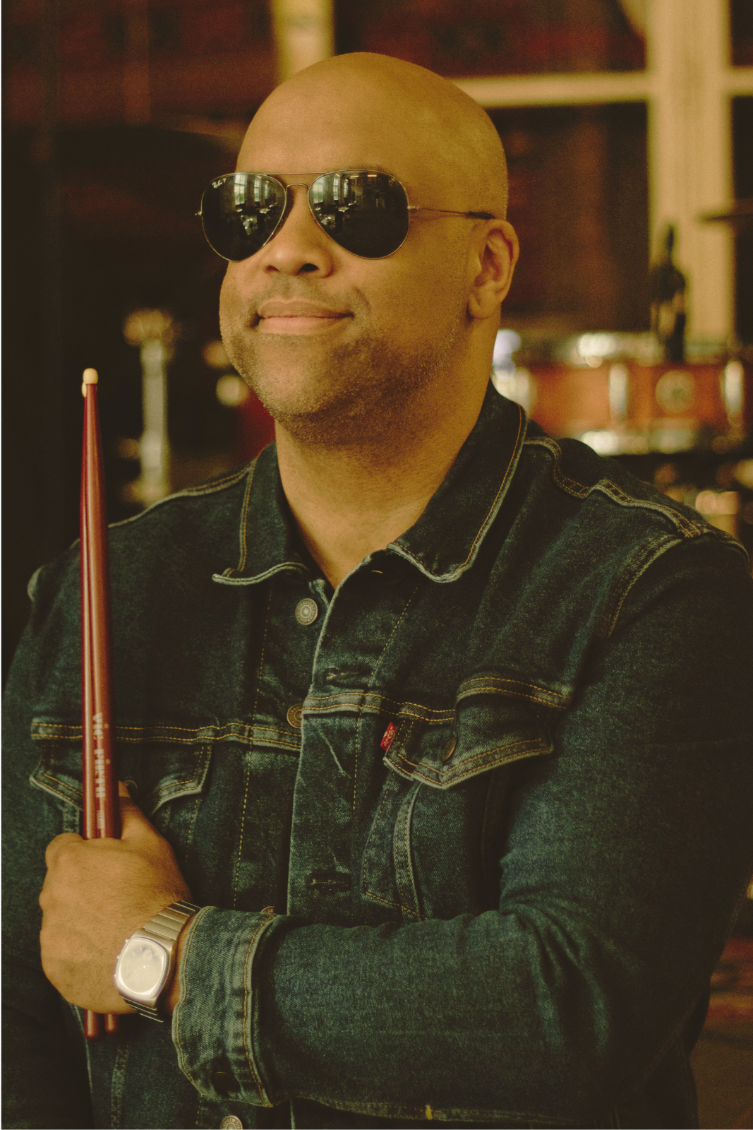 Mike Veny drummer and mental health speaker wearing sunglasses and a denim jacket, holding drumsticks in a music studio setting, representing resilience, creativity, and emotional expression.