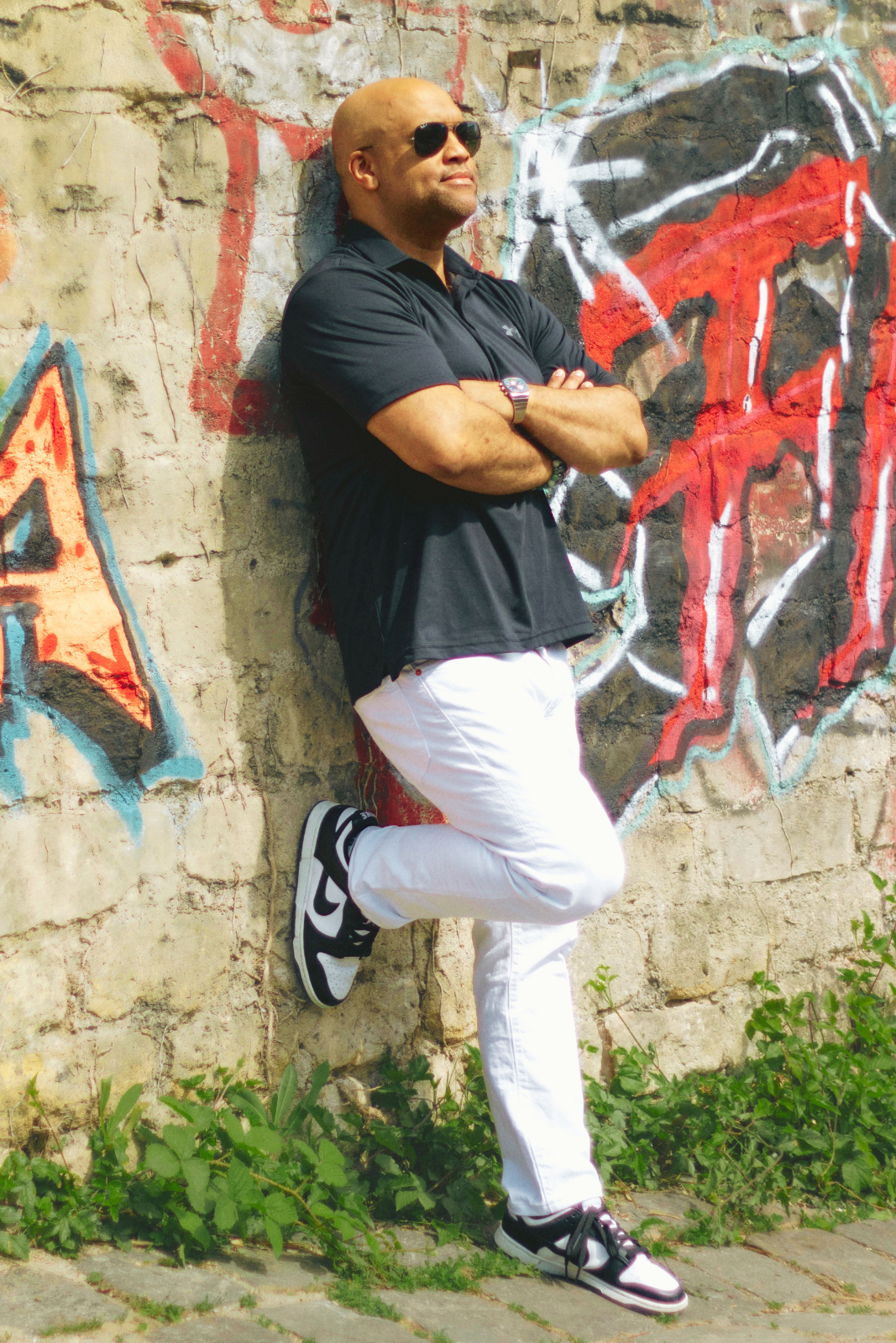Mike Veny drummer, speaker, and author leaning against a graffiti wall outdoors, wearing sunglasses and casual attire, representing confidence, creativity, and mental health advocacy.