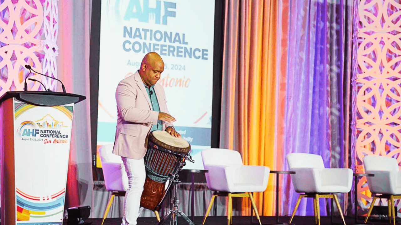 Mike Veny, healthcare keynote speaker and Certified Corporate Wellness Specialist, performs incorporated drumming during a keynote presentation at the Association for Healthcare Foodservice National Conference in San Antonio. He stands on a brightly lit conference stage beside a podium, white chairs, and colorful event lighting, creating an engaging emotional wellness experience for healthcare foodservice leaders.