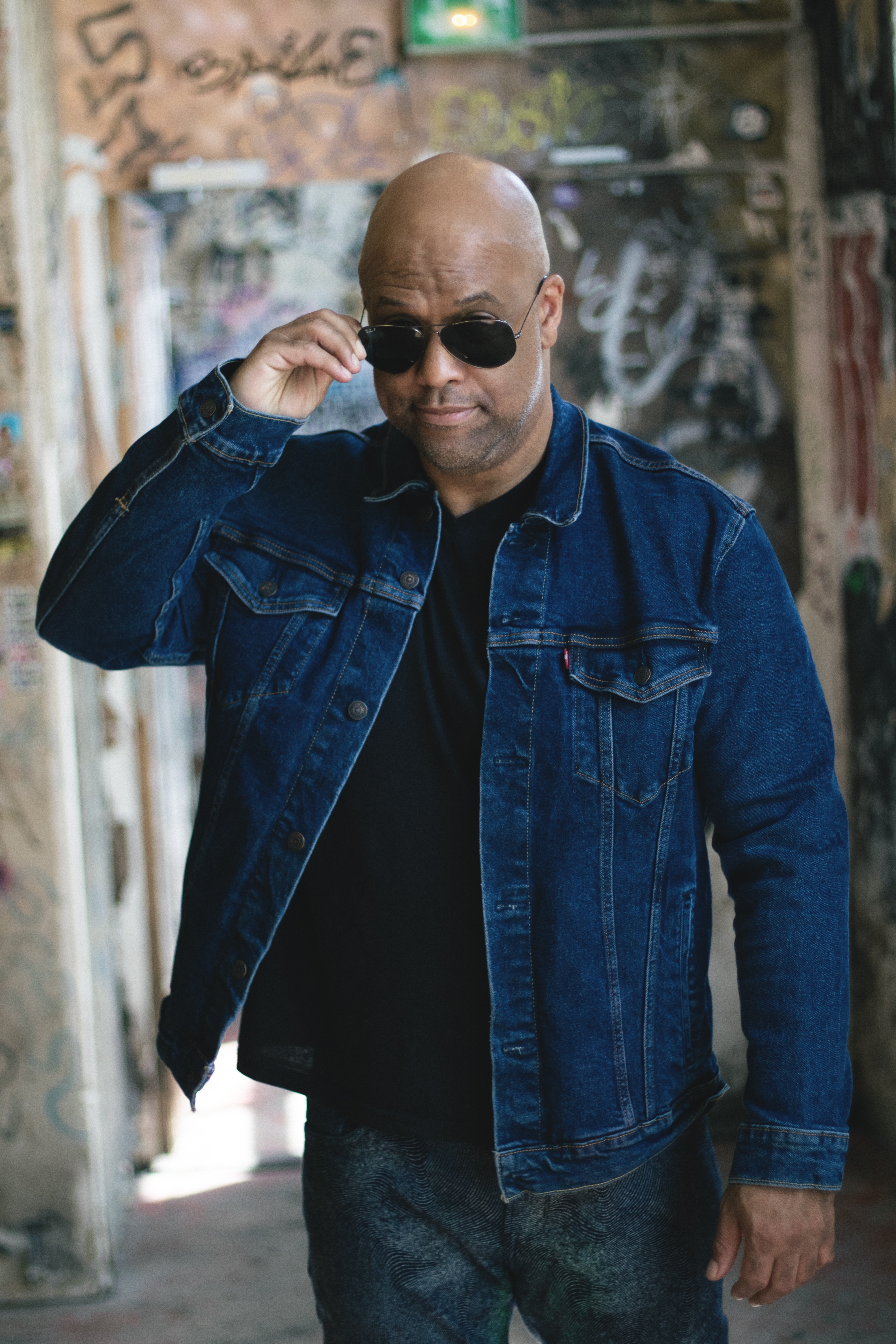 Healthcare speaker Mike Veny poses confidently in a denim jacket and sunglasses inside an urban graffiti-covered venue for mental health and workplace wellness event promotion.